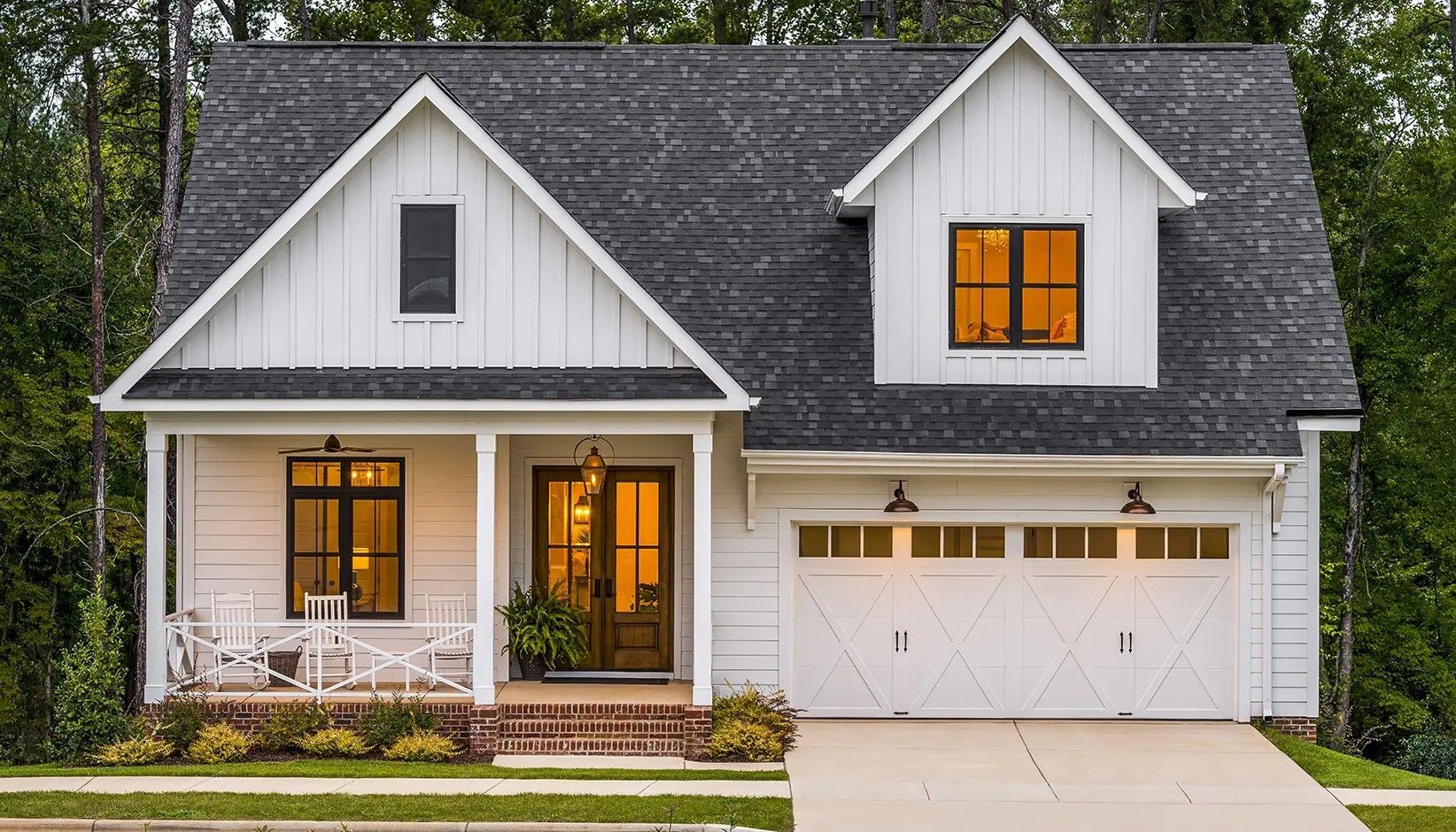 A white modern farmhouse with a dark shingled roof, a front porch, and a two-car garage, surrounded by green trees.