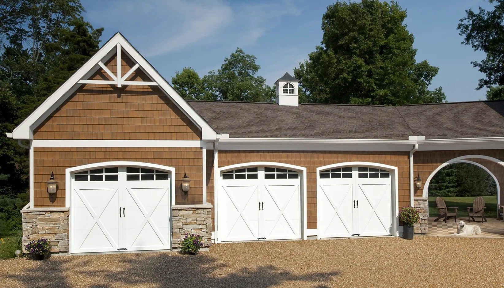 A brown, shingle-sided garage with three white carriage-style doors, stone accents, and an arched breezeway.