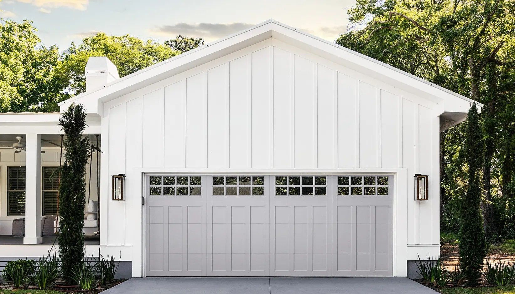 A white house with board-and-batten siding and a light gray garage door with windows, flanked by two tall, thin trees.