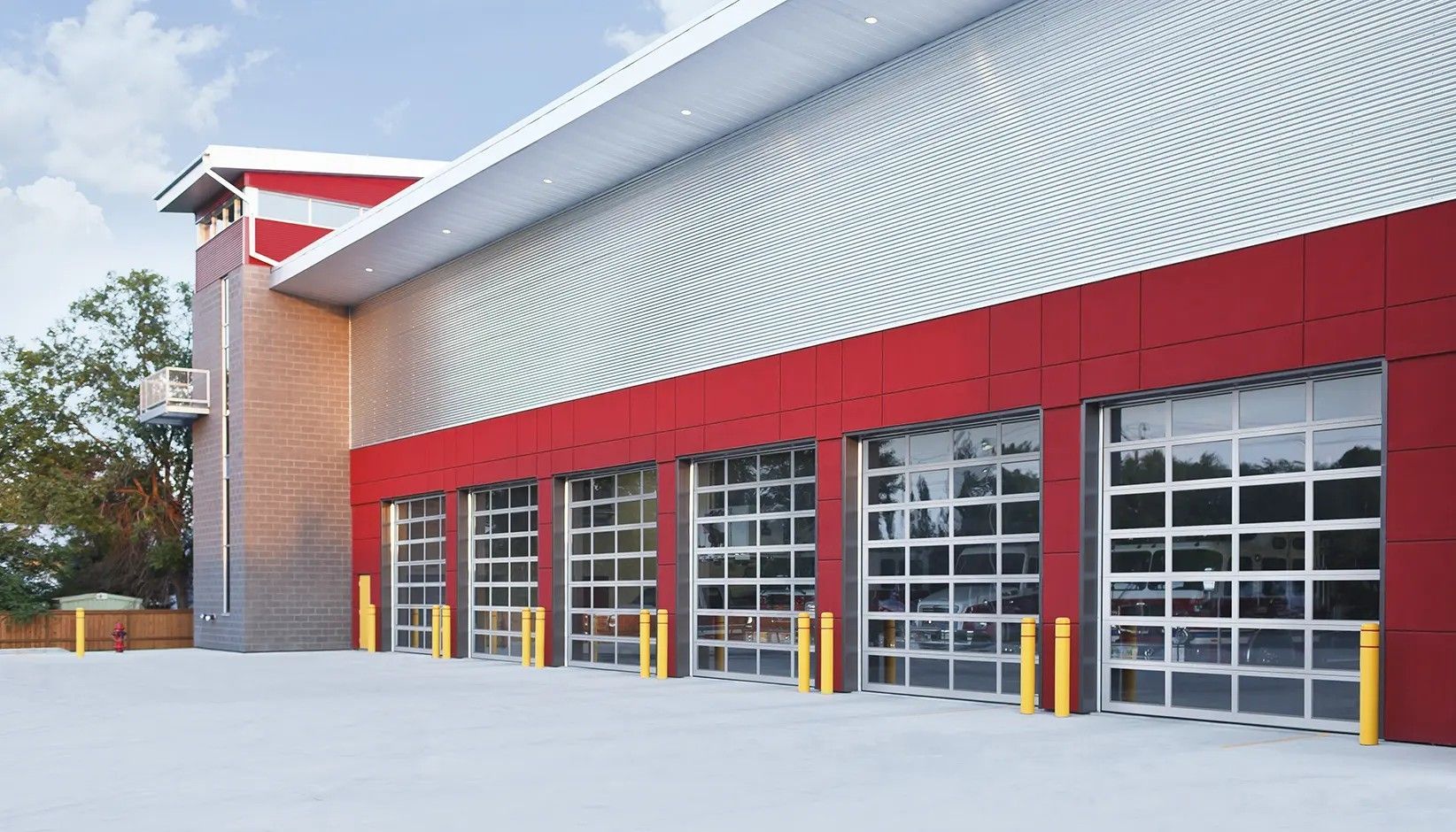Modern fire station with red walls and six glass bay doors, featuring yellow bollards on a concrete apron.