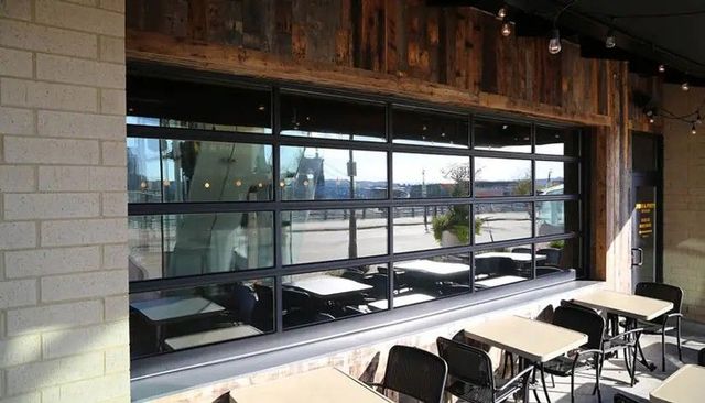 Outdoor patio dining area with tables and chairs next to a large, black-framed glass garage-style door on a brick building.