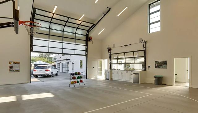 A modern, high-ceilinged indoor sports court featuring a basketball hoop, white walls, and a large roll-up glass door.