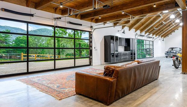 A spacious garage lounge with a brown leather sofa, patterned rug, glass garage doors, and a vintage car inside.