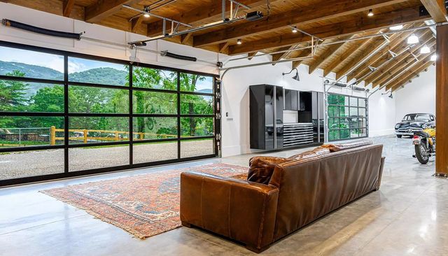 A spacious garage lounge with a brown leather sofa, patterned rug, glass garage doors, and a vintage car inside.