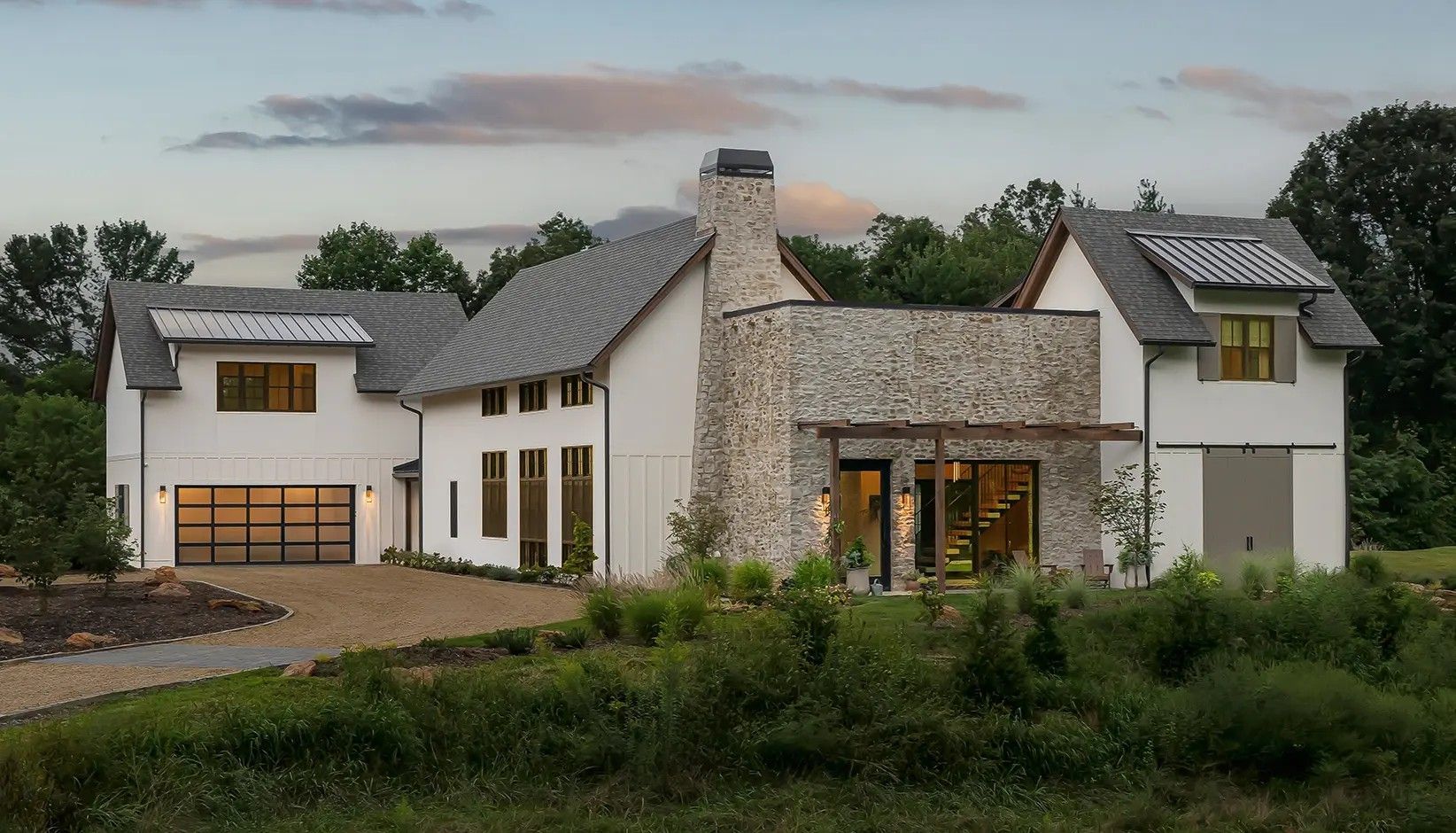 A multi-story modern farmhouse with white siding, stone accents, and a metal roof surrounded by trees at sunset.