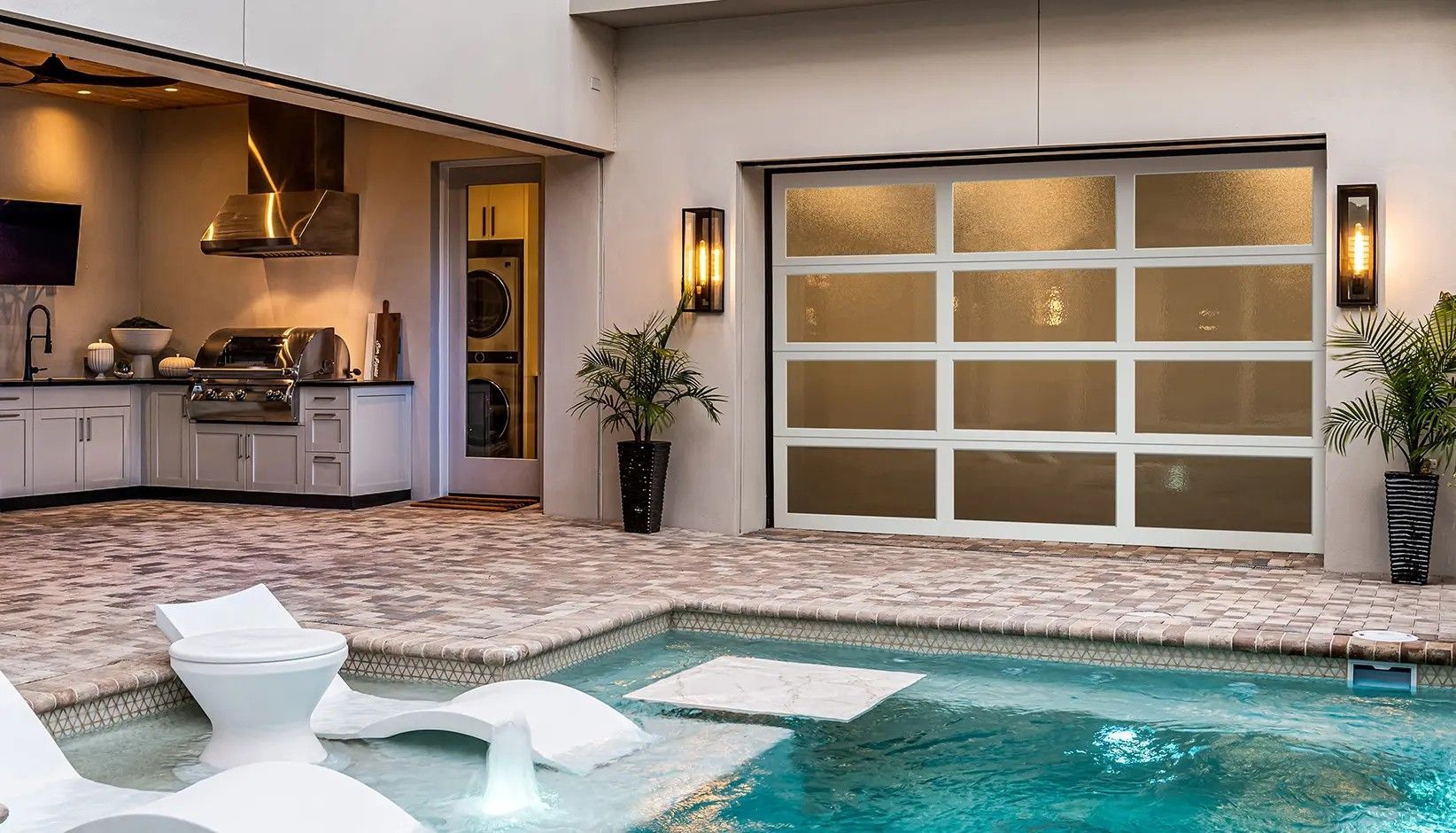 Modern outdoor kitchen with a pool, lounge chairs, and a glass-paneled garage door in a bright, patio setting.