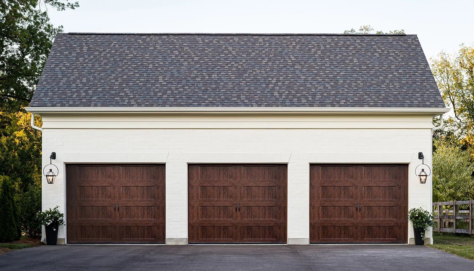 A three-car garage with white stucco walls, dark brown wooden doors, and a shingled roof, set against a tree-lined backdrop.