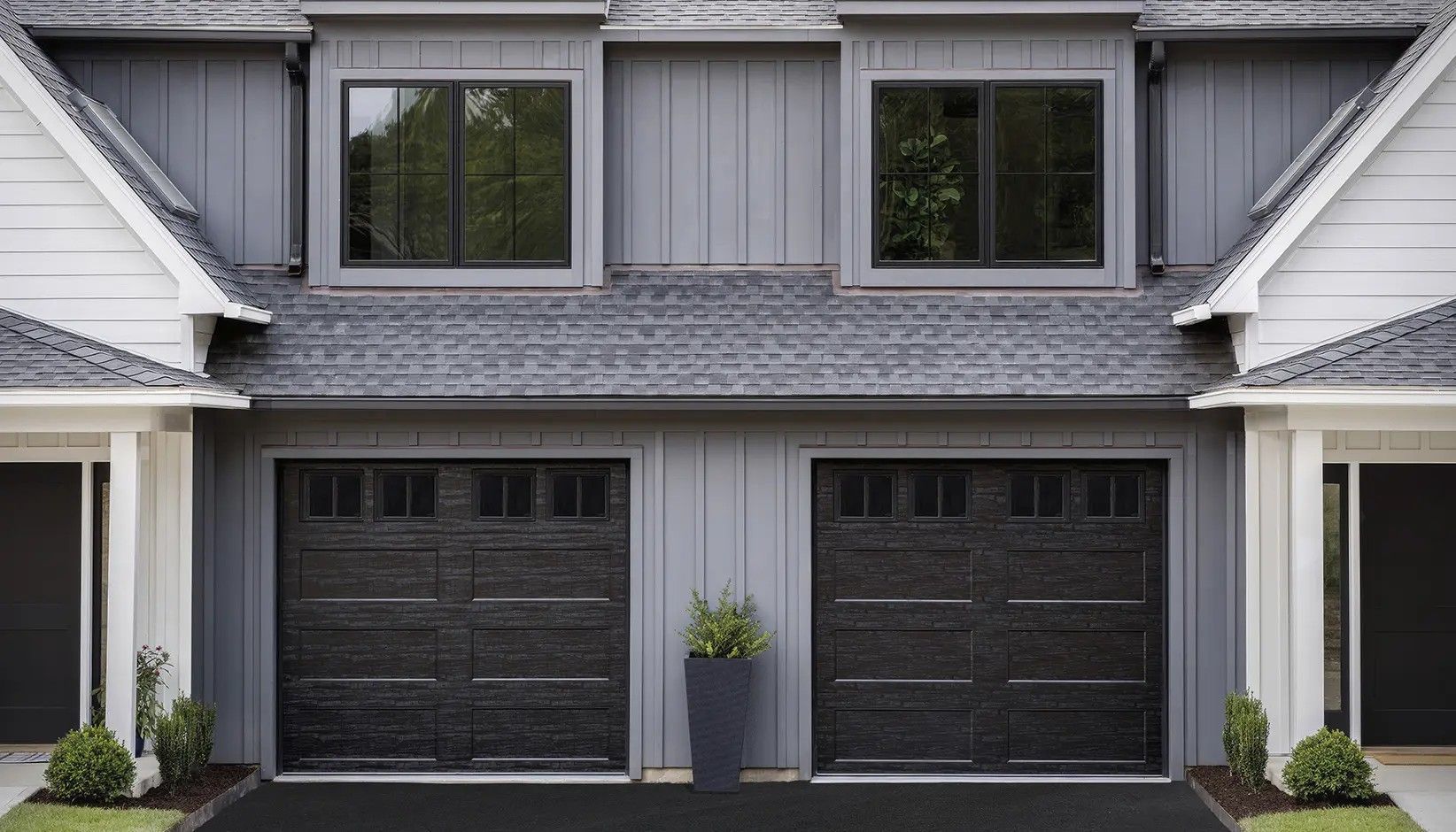 Two black garage doors on a grey home with a central potted plant, framed by white structures and two upper windows.