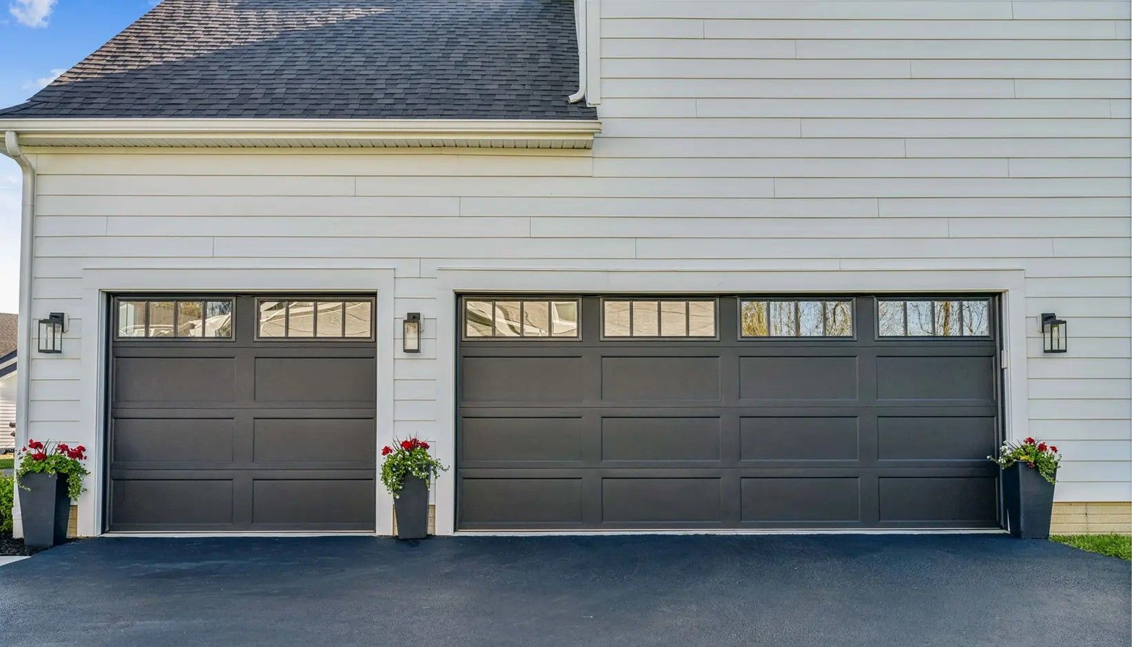 A white house with a two-car garage featuring charcoal-colored doors, window panels, and two potted red floral plants.