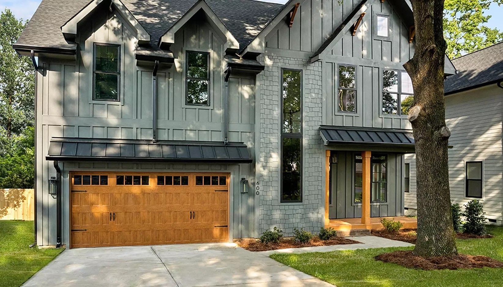 A two-story house featuring grey board-and-batten siding, a stone facade, a wooden garage door, and a metal roof.