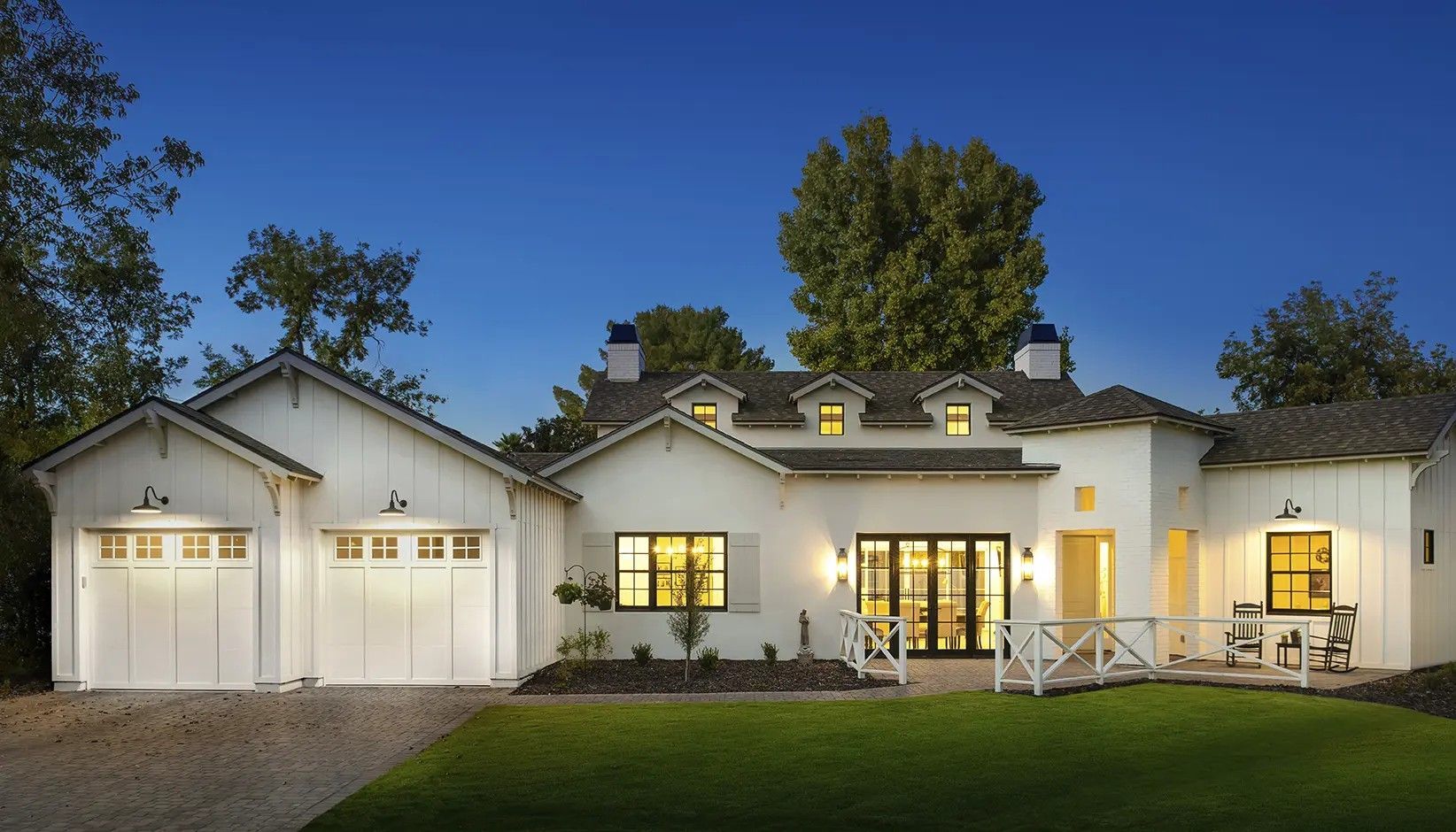A white, one-story modern farmhouse with a two-car garage at dusk, featuring a dark shingle roof and green lawn.