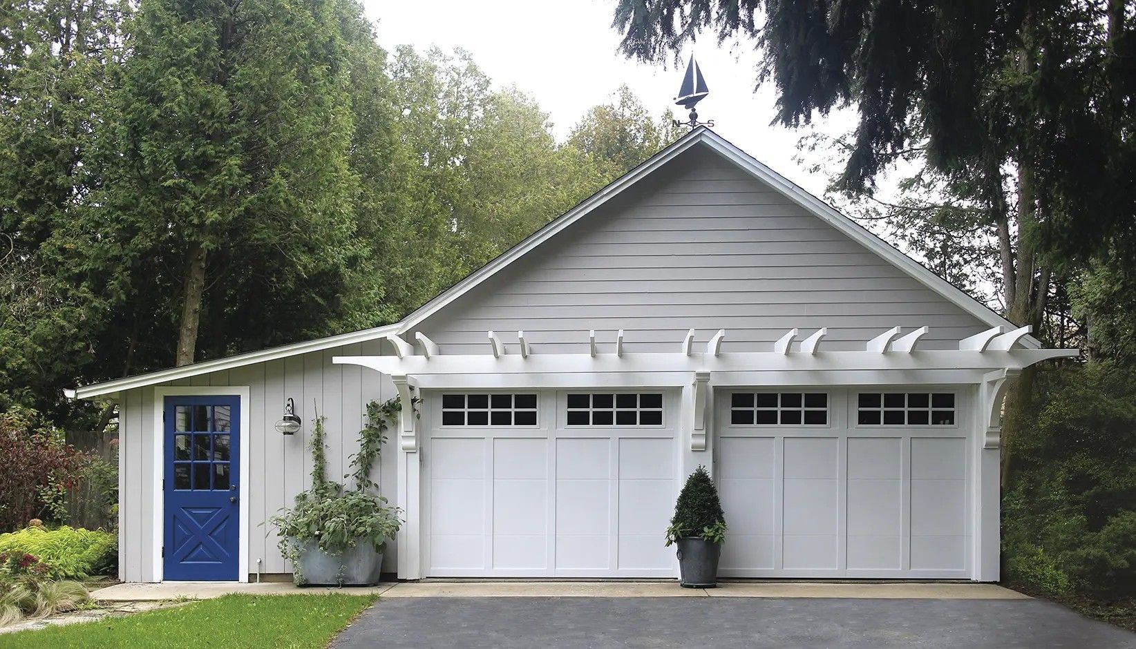 A gray detached two-car garage with white trim, a blue door, and a white trellis above the two garage doors.