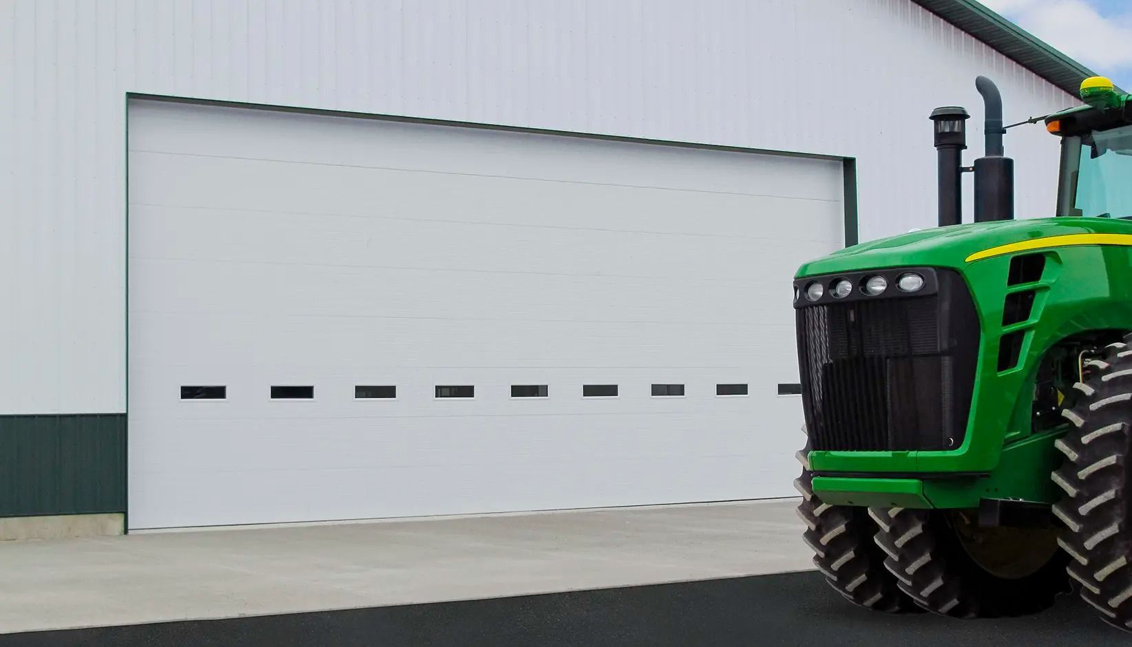 Green tractor parked beside a white garage door with small windows in an industrial yard