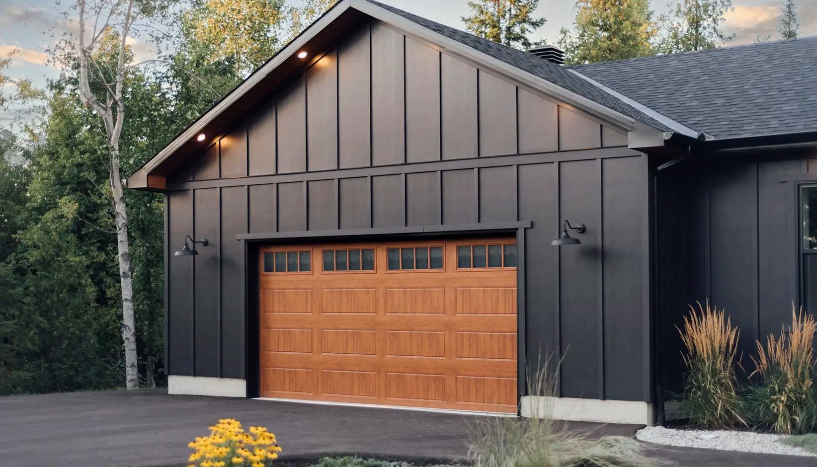 Modern dark-gray house with a wood-paneled garage door and lit exterior sconces in a landscaped driveway
