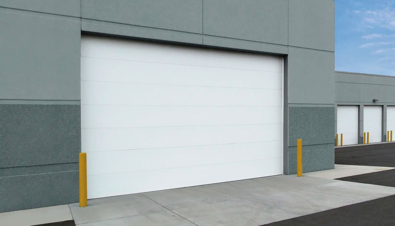 White industrial garage door on a gray building with yellow bollards beside a paved loading area