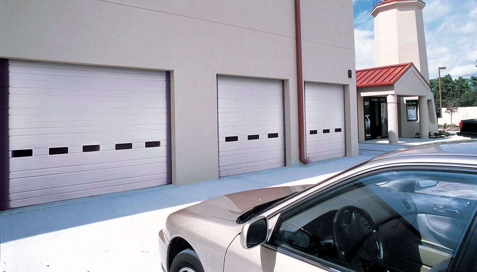 Row of white garage doors beside a beige building, with a silver car parked in the foreground and a red-roofed entryway.