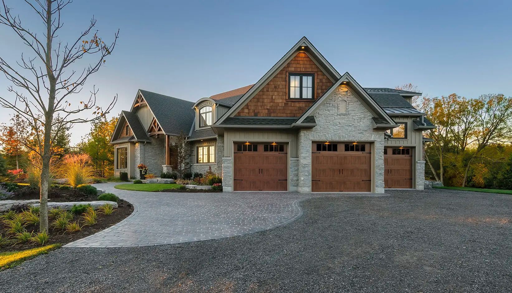 Modern stone house with three garage doors, gravel driveway, and autumn trees at sunset