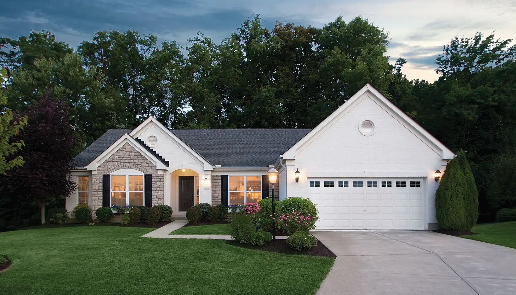 Suburban single-story house with white garage, lit porch, and landscaped front yard at dusk