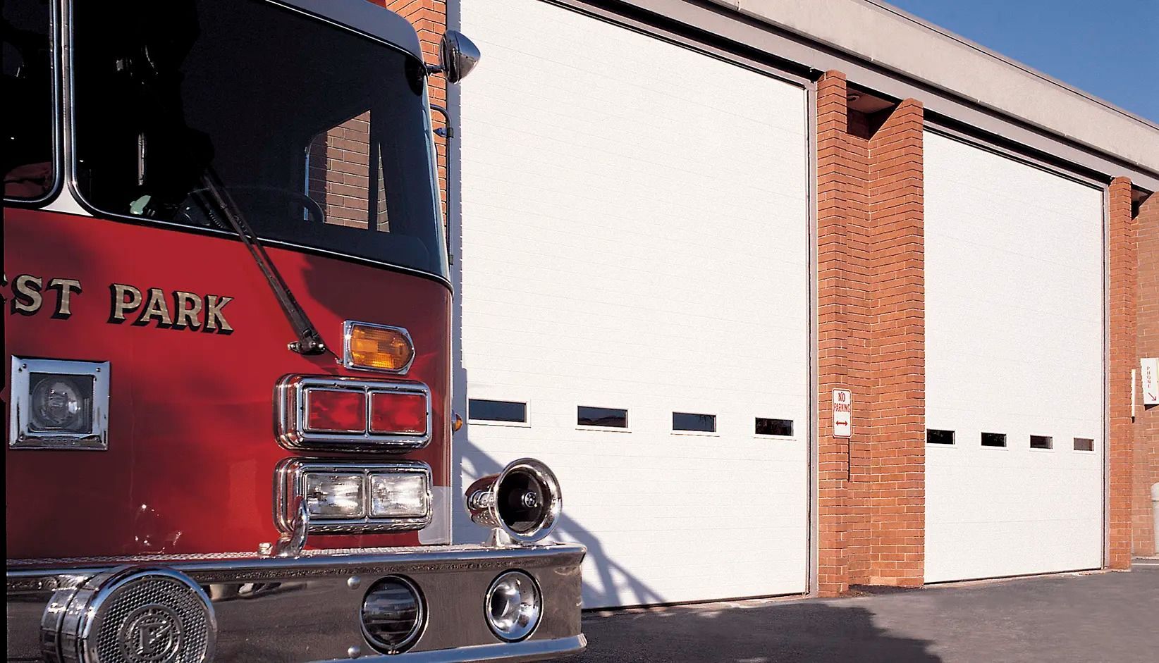 Red fire truck parked beside a station with white bay doors and brick columns