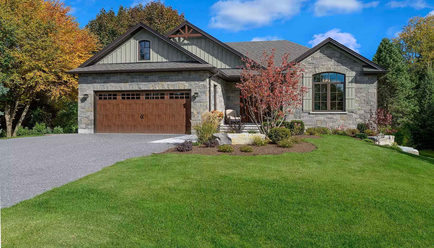 Suburban stone house with a two-car garage, green lawn, and autumn trees under a blue sky