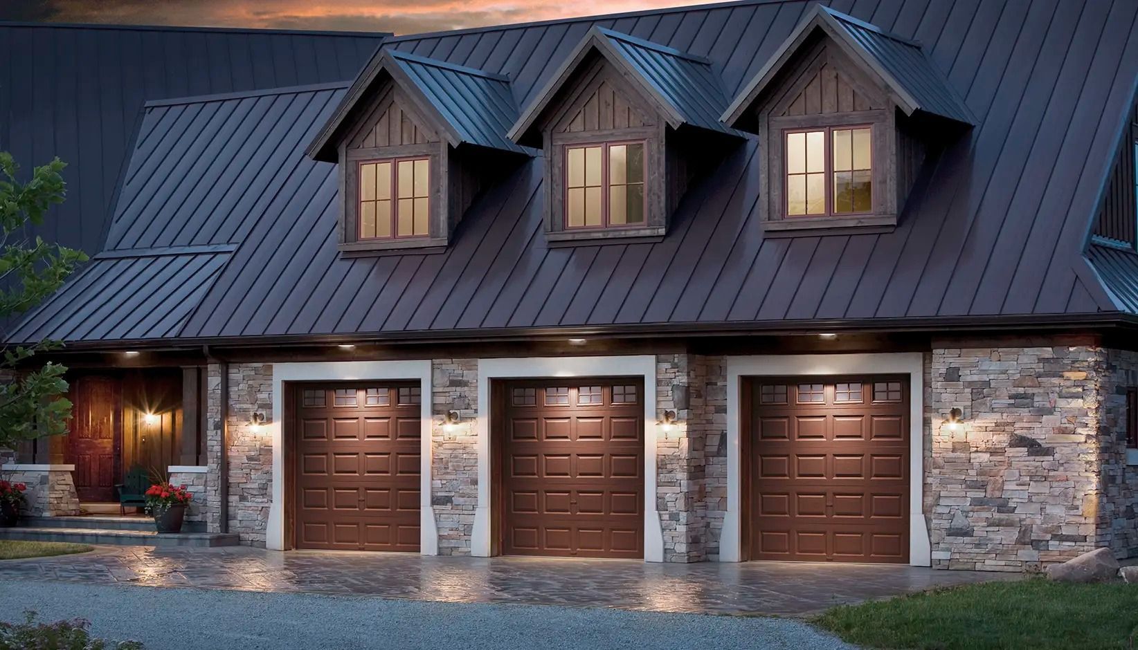 Stone-and-wood house exterior at dusk with three lit garage doors and dormer windows under a dark metal roof