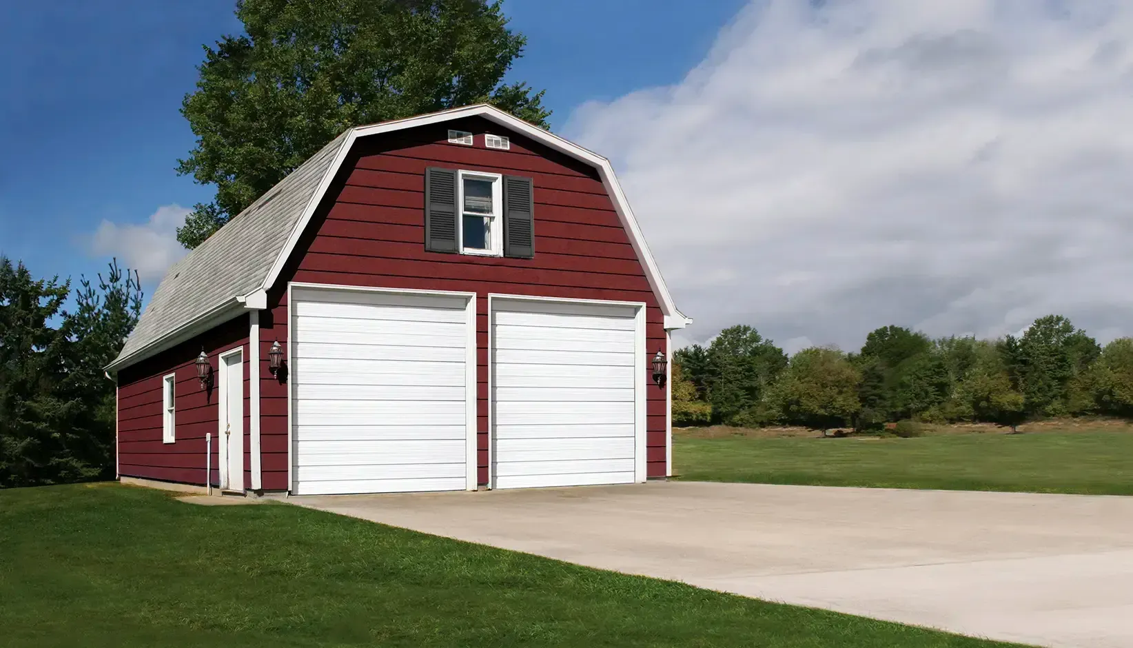 Red barn with white doors beside a driveway and green lawn under a blue sky