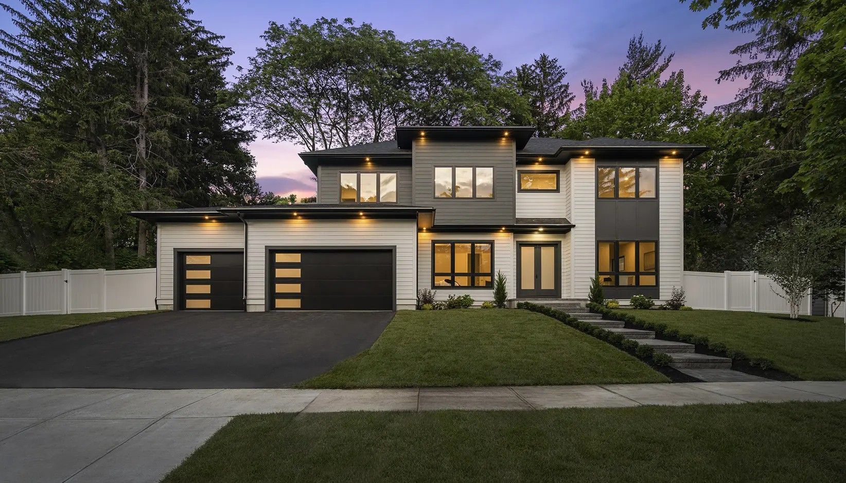 A modern, two-story house with a flat roof, dark accents, and two-car garage at twilight with landscaping.