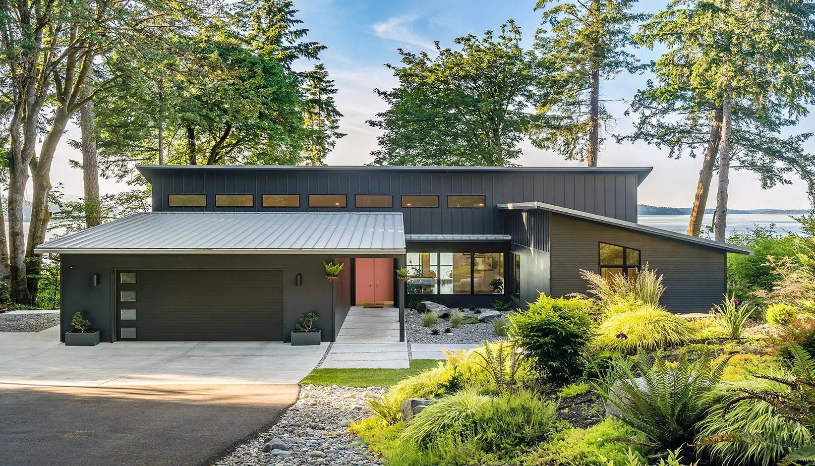 A modern, single-story gray house with a flat metal roof, a garage, a coral front door, and lush landscaping.