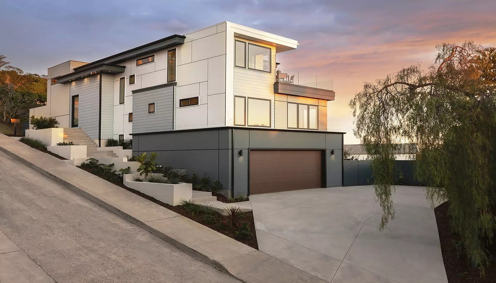A modern, multi-story house with a grey garage base and white upper levels, situated on a sloped lot at twilight.