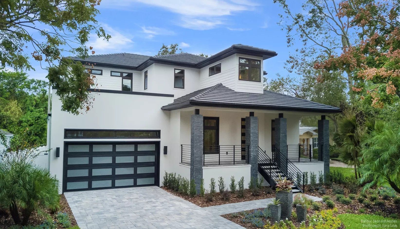 Modern two-story house with white stucco, a dark tile roof, a glass-paneled garage door, and a stone-pillared porch.