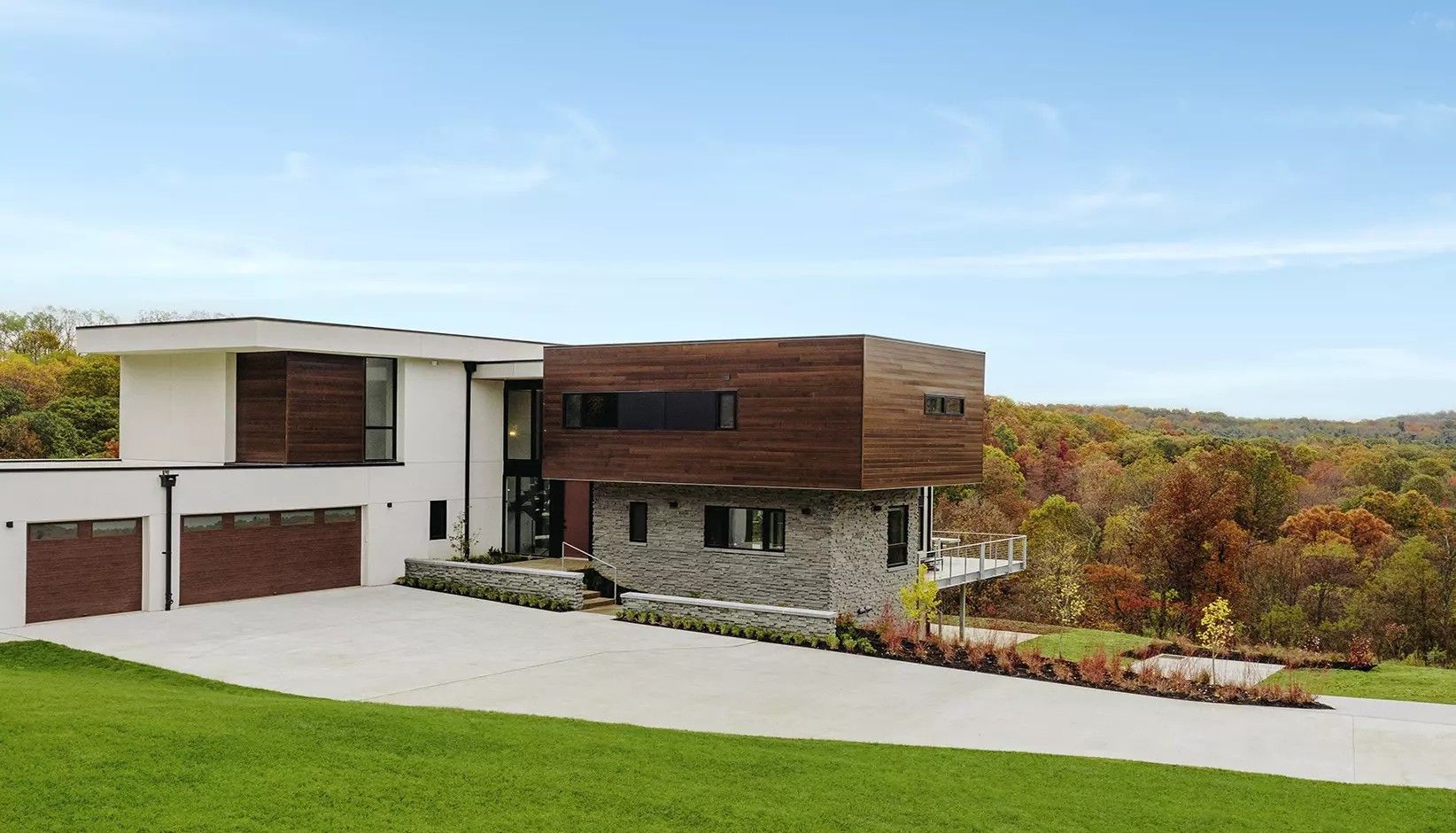 A modern house with white, wood-paneled, and stone sections, featuring a large driveway, set against a wooded landscape.