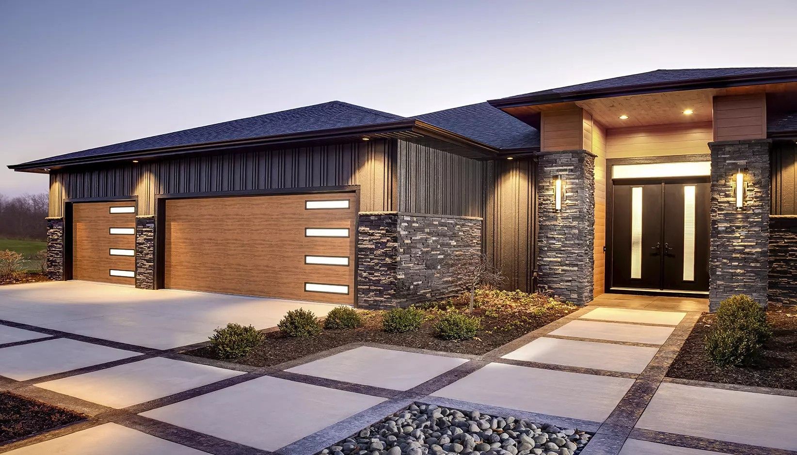 Modern one-story home at twilight with a three-car garage, stone masonry accents, wood siding, and a lit concrete path.