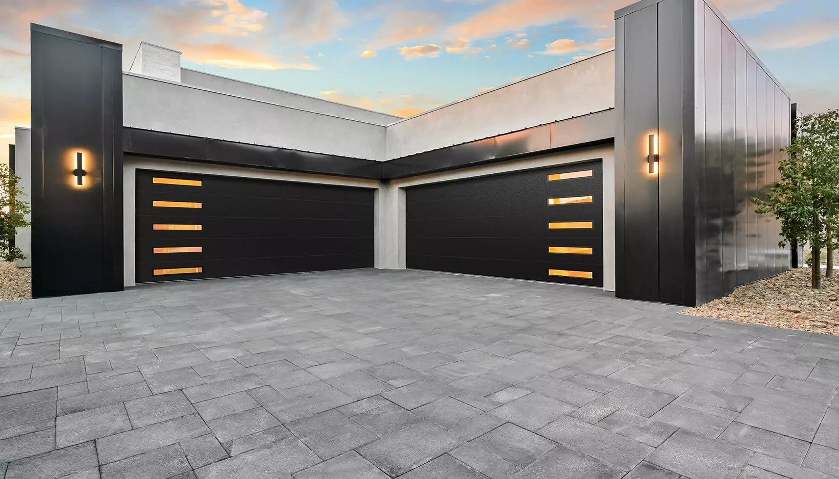 Modern house exterior featuring two black garage doors with horizontal glass windows, stone driveway, and glowing sconces.