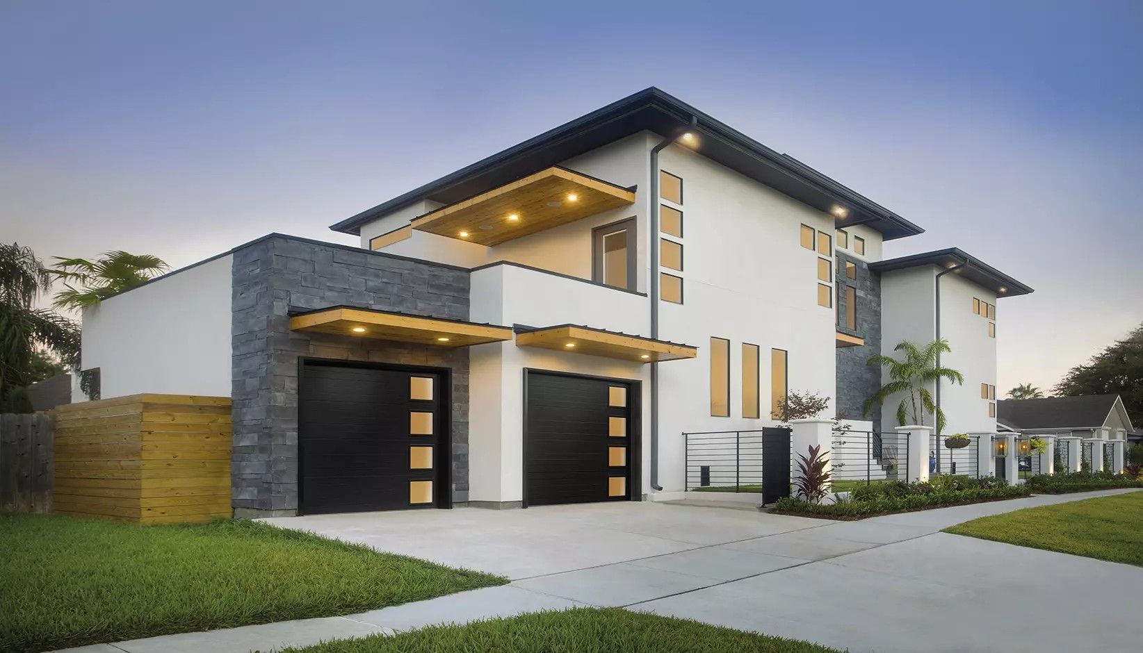 A modern two-story home with white stucco, dark stone accents, and two black garage doors under wood-toned overhangs.