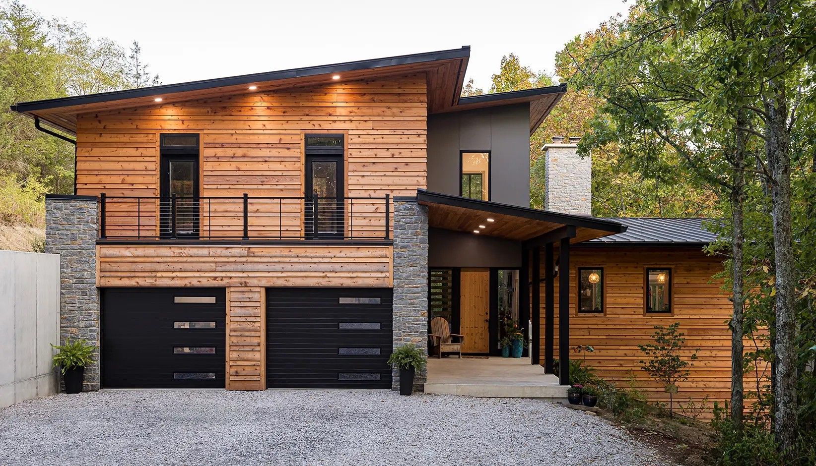 A modern two-story home with wood siding, stone accents, a shed roof, and two black garage doors set in a wooded area.