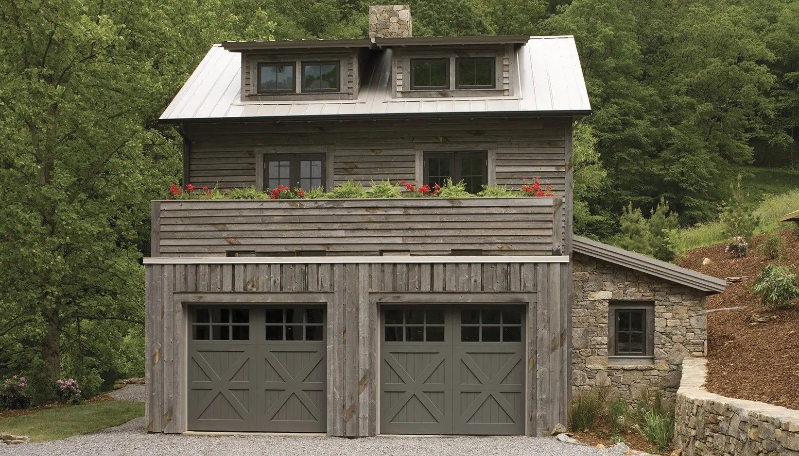 A rustic two-story wooden carriage house with a stone side room, two dark grey garage doors, and a flower-filled balcony.