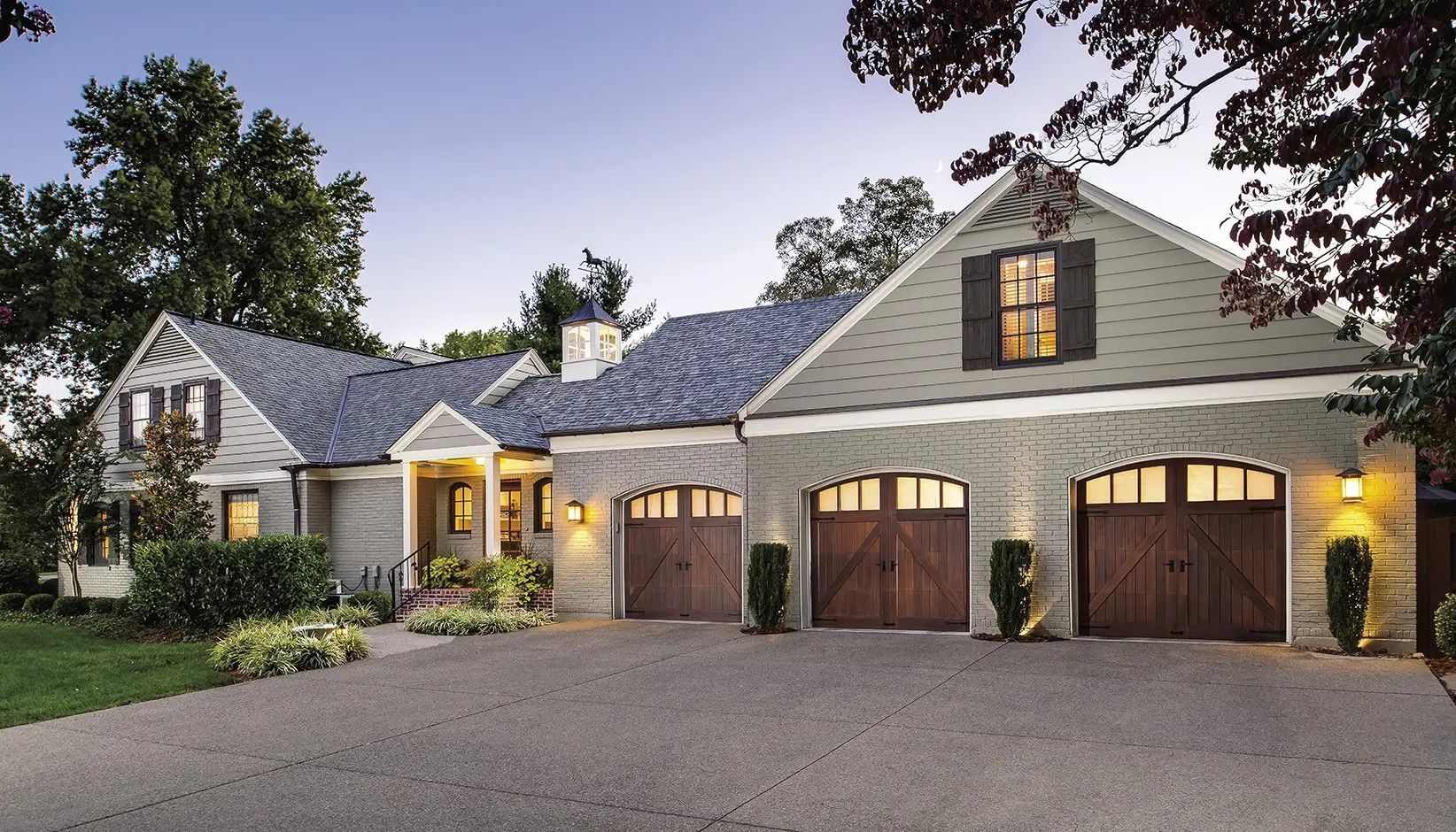 A sprawling, light-colored house with a three-car garage, multiple gabled roofs, and a gravel driveway at dusk.