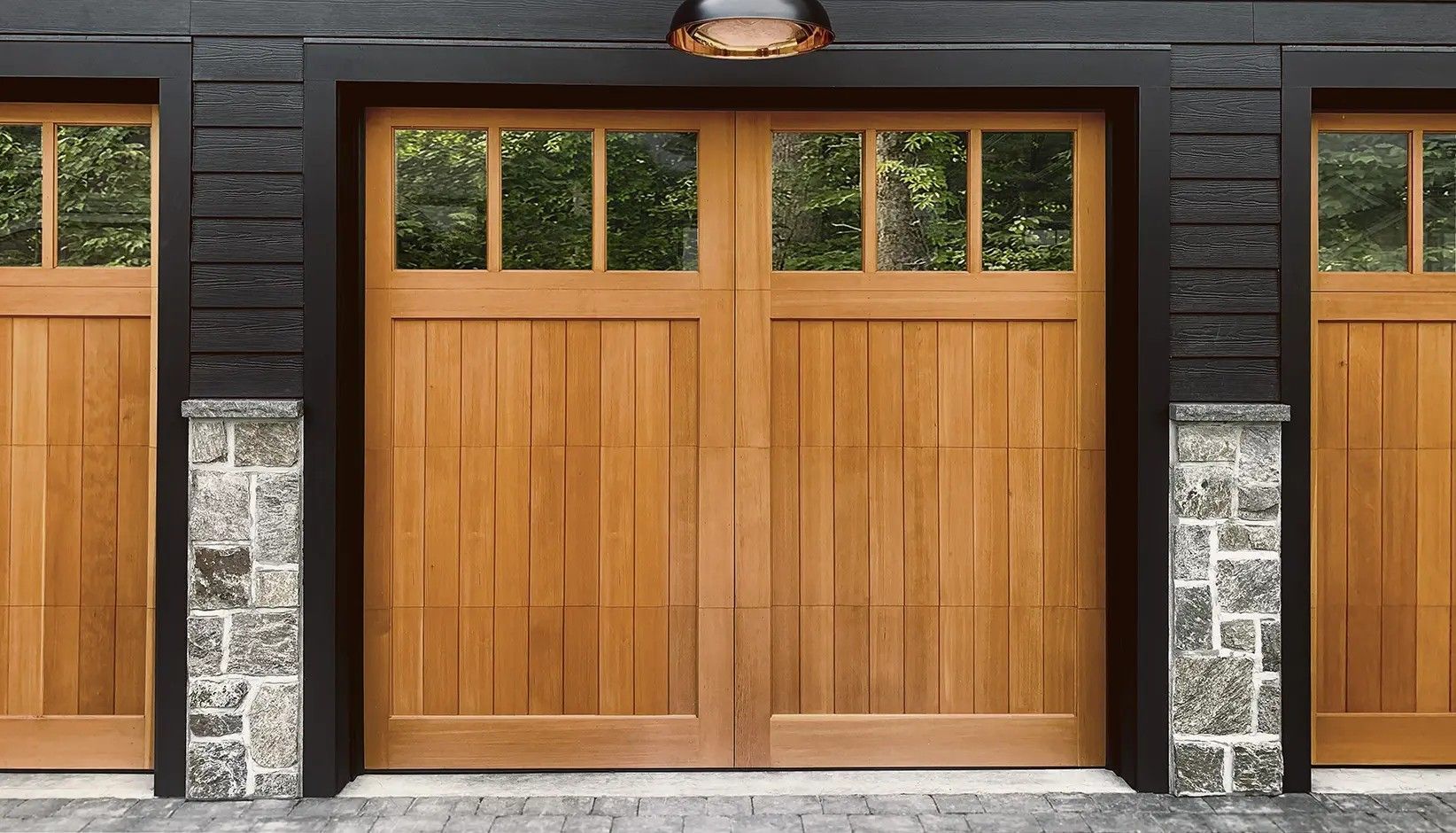 Three wooden garage doors with glass window panels set against a dark frame and stone pillars.