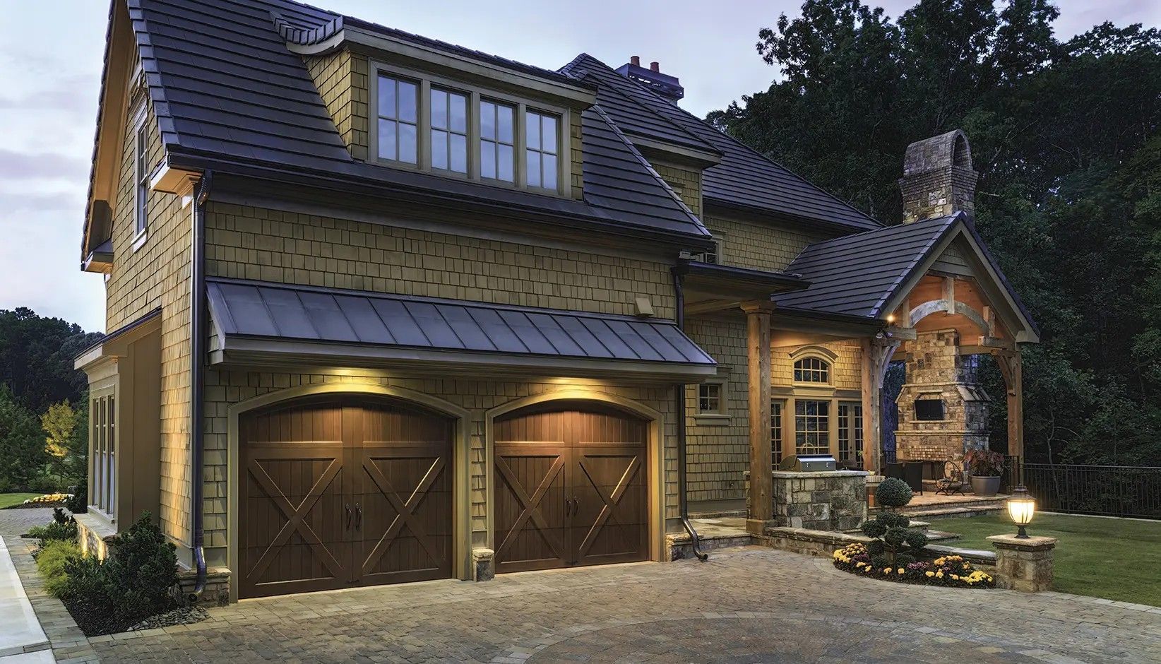 A two-story stone home with a dark shingle roof, double garage doors, and an outdoor stone fireplace at dusk.