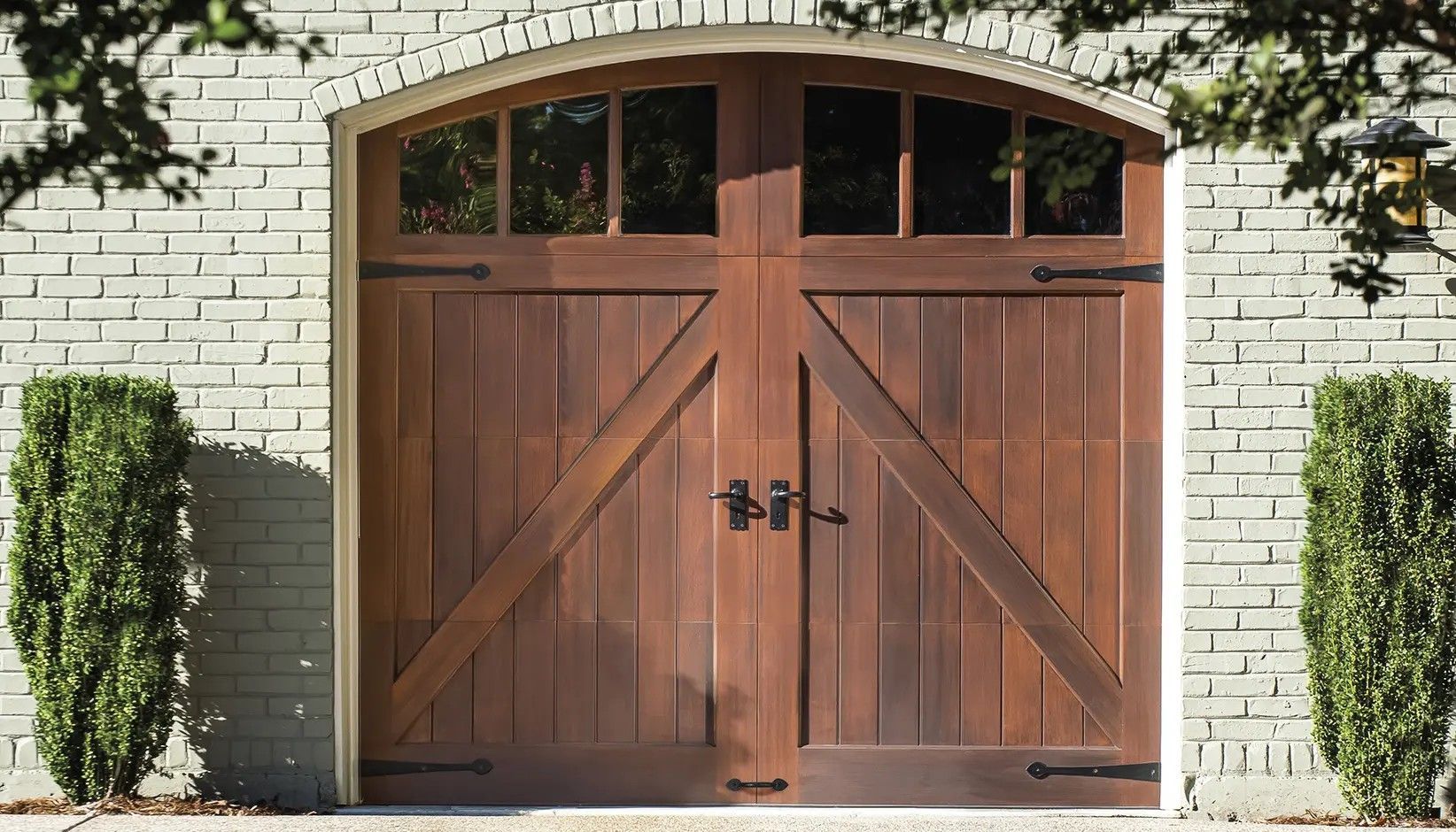 Arched, wood-toned carriage-style garage door with windows, decorative black hinges, and hardware set in a brick wall.
