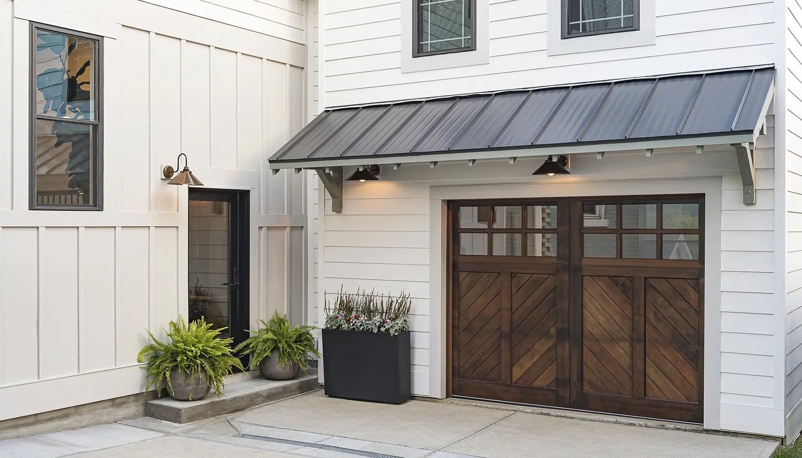 A white home exterior with a dark-roofed garage, wood carriage doors, a side entry door, and potted greenery.