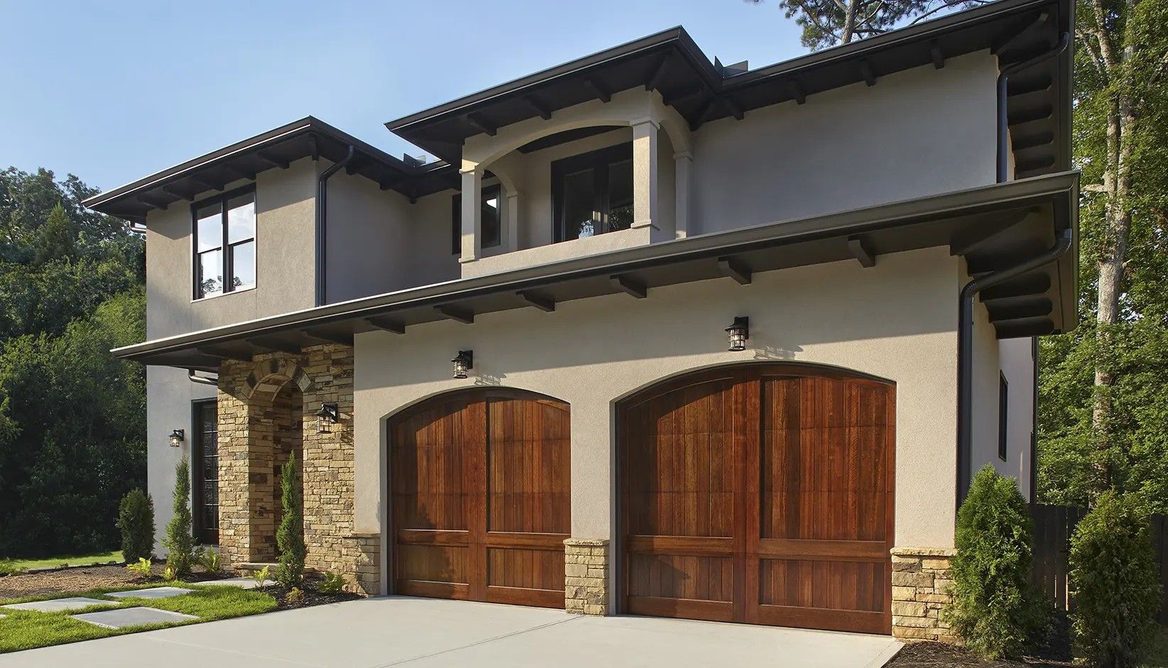 A two-story stucco house with a stone facade, dark wood arched double garage doors, and a matching overhanging roof.