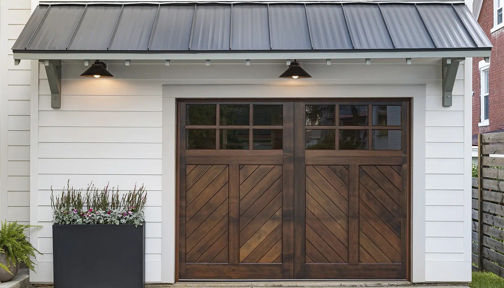 A dark wood garage door with a diagonal slat pattern, set in a white-sided building with a black metal awning and lights.