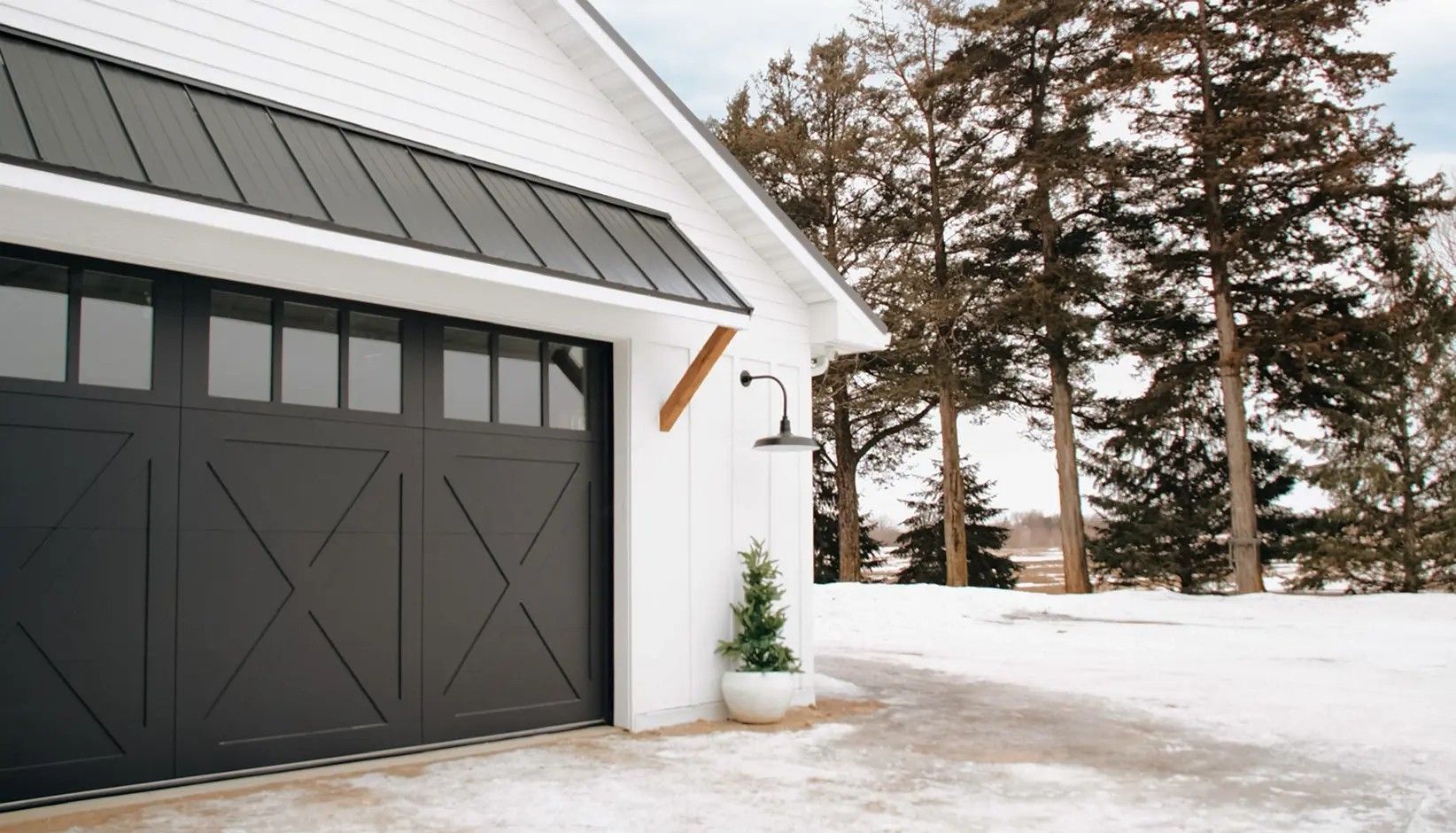 Black garage door with X-pattern and windowed top panel on a white building, set in a snowy landscape.