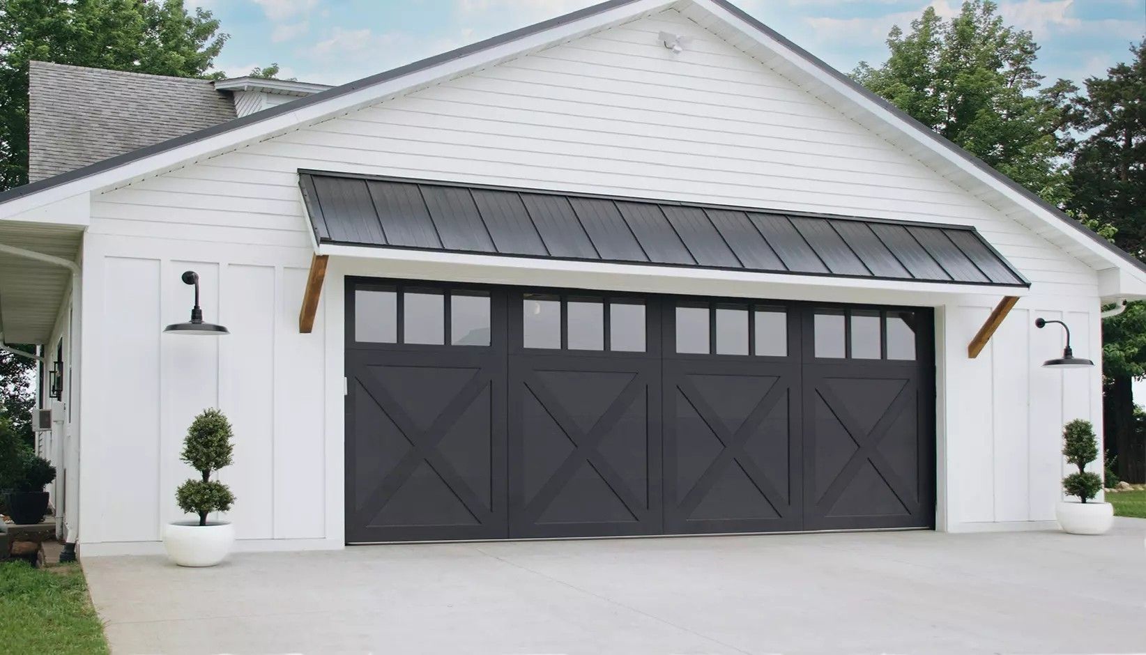 A white garage with a black carriage-style door, a metal awning, and exterior wall lanterns on a concrete driveway.