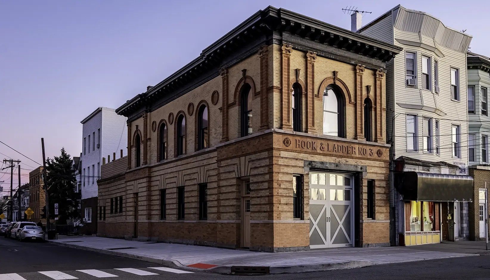 A two-story tan brick building with arched windows and a white garage door on a city corner at dusk.