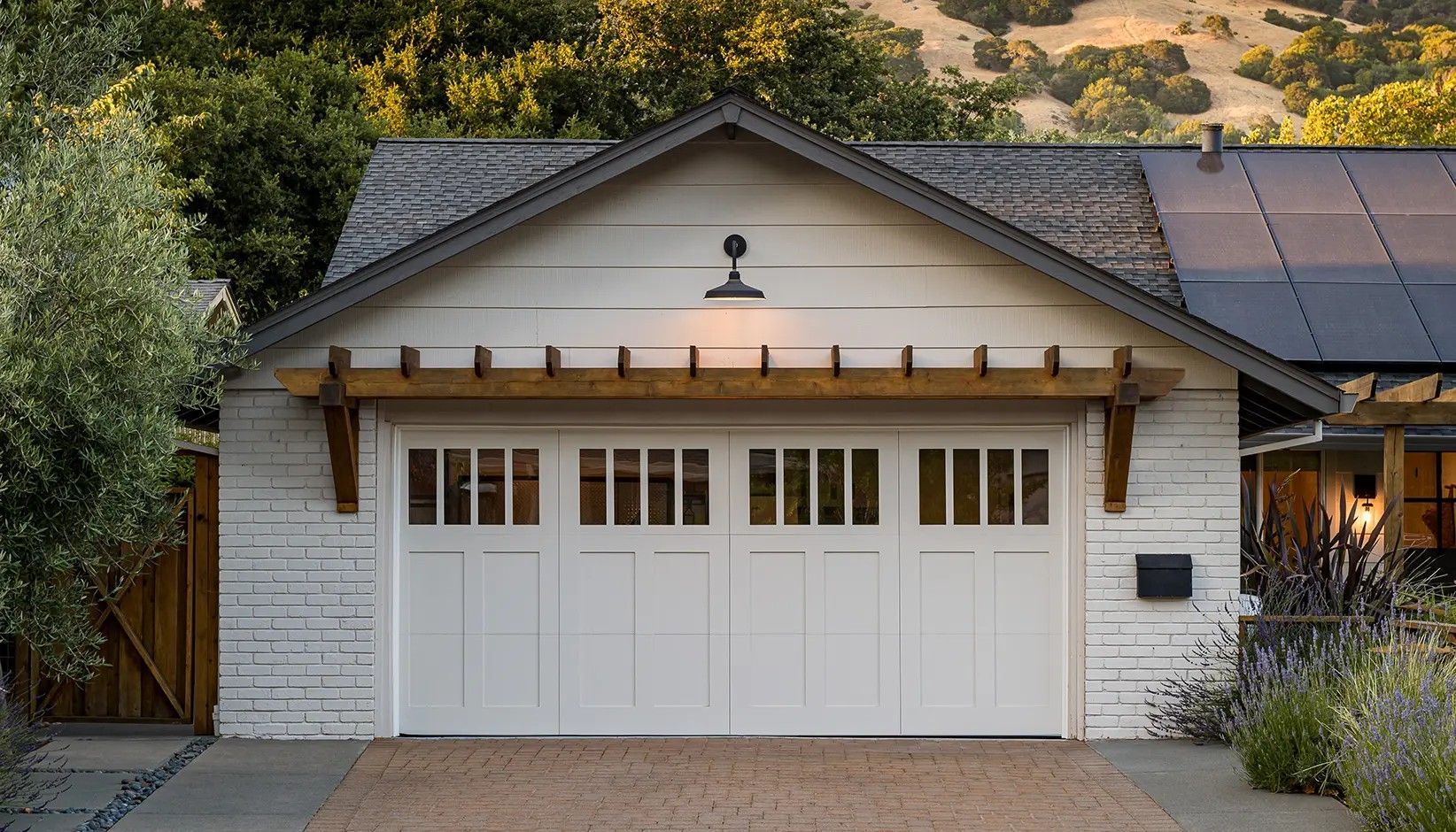 A white, detached garage with a wooden pergola, windowed door, and exterior light against a backdrop of trees.