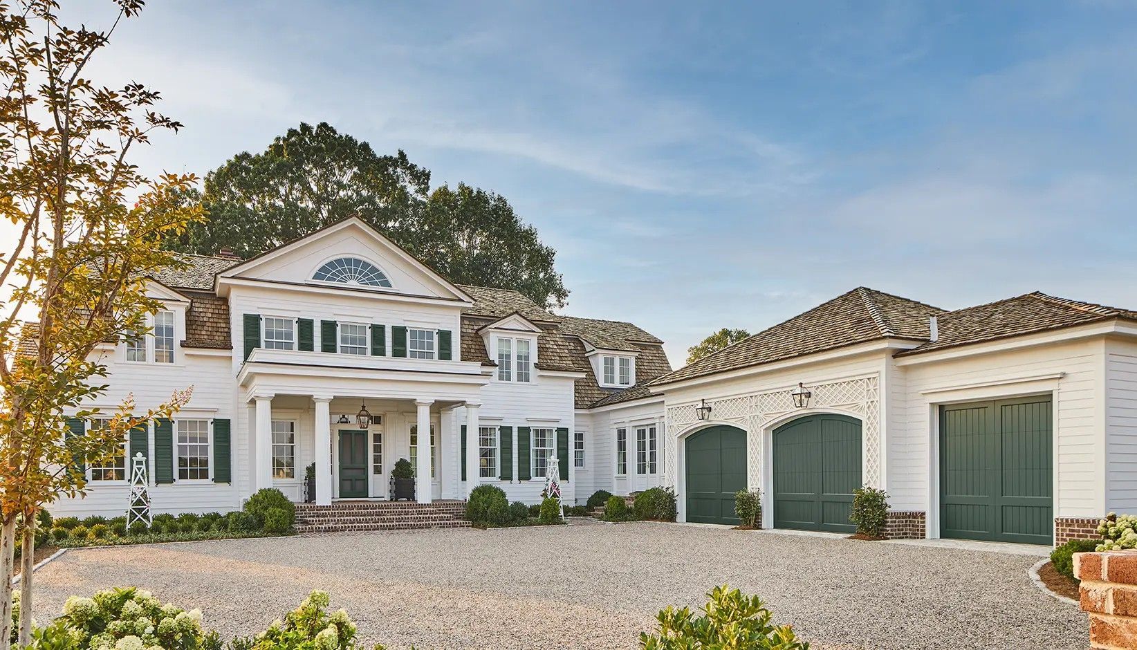 A large, white two-story colonial-style house with green shutters, a gravel driveway, and a three-car garage.