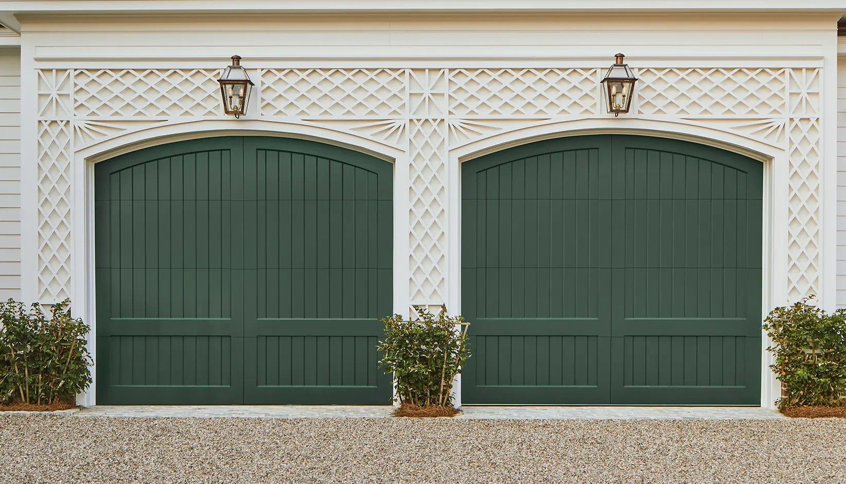 Two arched, dark green wooden garage doors framed by decorative white lattice trim, featuring wall lanterns above each.