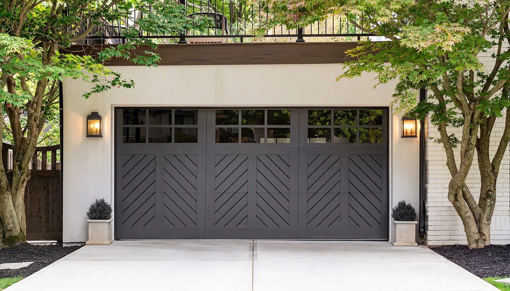 A charcoal-colored garage door with a herringbone pattern and rectangular windows, set between two trees and wall lamps.